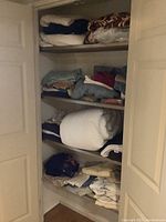 Closet shelves showing stacks of folded used linens including blankets, towels, and bedding items arranged across four wooden shelves.