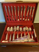 Opened wooden box with upper section holding 12 knives secured by felt loops and lower section holding 6 large spoons, 6 large forks, 6 medium spoons, 6 medium forks, plus additional loose pieces in a plastic bag.