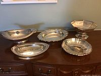 Five silverplated serving pieces on a wooden sideboard, showing overall shapes and size