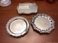 Photo of three silverplated serving trays on a wooden table, showing oblong tray with pierced sides and two round trays with decorative edges and engraved surfaces.