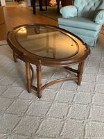 Oval solid wood coffee table with glass top positioned on a carpeted floor next to a blue chair.