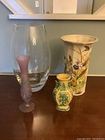 Four assorted vases on a wooden surface. One large clear glass vase, one small purple frosted glass vase, one small ceramic marked from Poland, and one taller ceramic vase painted with flowers and butterflies.