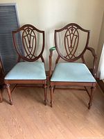Two vintage wooden dining chairs side by side on hardwood floor against a light wall, one armless and one with wooden arms, both with shield-shaped carved backs and light blue upholstered seats.
