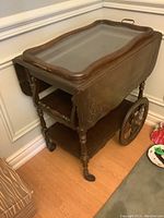 Side view of vintage wood tea cart showing removable glass serving tray, drop leaf extended, wheels and turned legs.