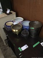 Wood bench with assorted metal and porcelain planters displayed on top, showing tarnish on metal items and varying colors and styles of planters.