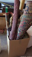Multiple rolled carpets in a cardboard box showing various traditional rug patterns and colors including red, beige, and purple designs.