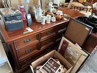 Brown wooden 10-drawer dresser with various decorative items on top and a box with miscellaneous papers in front.