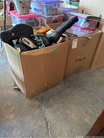 Two large cardboard boxes filled with various shoes and boots, some with visible soles and laces, along with smaller bins on top and nearby indicating shoe care items.