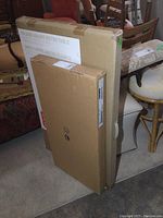 Flat packed IKEA Lillången cabinet box and folding bistro table boxed and leaning against furniture.