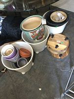 Grouping of various planters stacked together on concrete floor including white porcelain planters, a colorful floral and bird design planter, and terracotta small pots alongside wooden IKEA plant dollies.