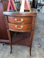 Front view of vintage wooden side table showing two small curved drawers with brass handles and floral carved wooden sides.