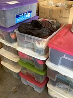 Multiple plastic storage bins stacked containing scarves, hats, gloves, belts, and a wig as women's accessories.