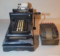 Two adding machines side by side: large Burroughs machine on left with keys and paper roll, smaller AMCO machine on right with rotary dials and red buttons.