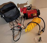 Air compressor with yellow coiled hose, battery clip cables and carrying bag laid on surface.