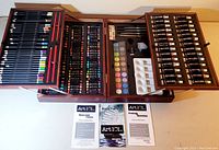Full view of open wooden art case with all compartments showing colored pencils, oil pastels, acrylic paint tubes, watercolor cakes, and instructional pamphlets laid out.