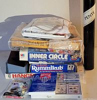 Stack of games including Michigan Rummy, Inner Circle, Rummikub, Hangman with cards, dice, dominoes nearby and wine bottle for scale