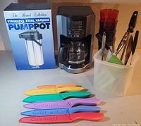 Photo showing coffee maker, boxed stainless steel vacuum pump pot, colorful sheathed knives, and container with assorted kitchen utensils on countertop
