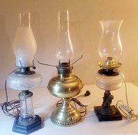 Photo showing all three antique oil lamps converted to electric lighting. From left: clear glass base lamp with frosted white font and chimney; brass base lamp with clear chimney; white ceramic lamp with cherub base and clear chimney.