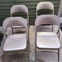 Four beige metal folding chairs arranged side by side outdoors on concrete, showing condition and style.