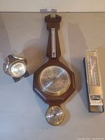 Full view of all three items: the wooden weather station, the multi-sided barometer/thermometer unit, and the packaged outdoor thermometer.
