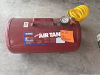 Red Air Works portable air tank with pressure gauge and yellow coiled hose visible on top, placed on a concrete floor