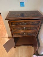 Front view of wooden cabinet showing top surface, three drawers with decorative carvings and metal knobs, and lower door storage compartment open.