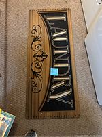 Photo of one wooden laundry mat with curved 'LAUNDRY' lettering and ornamental black scroll design, seen on carpeted floor next to washer and dryer.