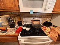 Full view of kitchen counter with stacked glass pie pans, white bowls, wooden knives in block, plastic containers, and utensils laid out near stovetop.