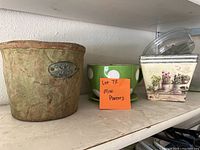 Photo showing three planters on a shelf: a textured ceramic planter with a metal plaque, a green planter with white polka dots, and a cream-colored planter with floral images and handles.