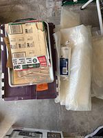 Photo showing stacked boxes, sheets of 3M sandpaper, plastic drop cloths, and paint brushes in a plastic container.