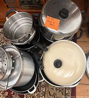 Top view of a stack of various pots and pans including stainless steel pots with inserts and black handled pans, with an off-white enameled pot on top.