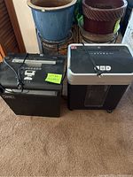 Two paper shredders side by side on carpet in front of potted plants, one black Fellowes and one black and silver Ativa shredder.