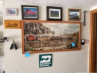 Several framed train pictures and signs on a wall including a large framed train landscape print, smaller framed train photos, a small wooden train sign, and a Burlington Northern sign below them.