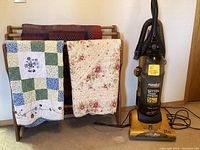 Photo showing Eureka vacuum cleaner next to a wooden quilt holder stand draped with three quilts of different floral and patchwork designs.