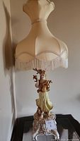 Full view of the antique lamp showing fabric shade with fringe and lady figurine base on ornate pedestal.