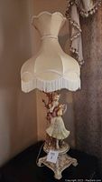 Full view of one antique table lamp with lady figurine base and fabric lampshade with long white fringe