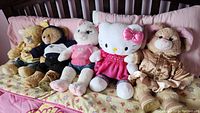 Five plush toys lined up on a bed including a yellow teddy in jacket, a brown bear in white t-shirt, a white cat, Hello Kitty in pink dress, and a beige bear in pink satin jacket