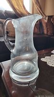 Clear glass water pitcher photographed from a front angle showing curved handle, wide base, and spout.