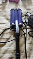 Three-barrel purple and black hair crimper laid on beige fabric surface, showing handle and barrel plates with attached power cord.