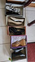Five pairs of ladies shoes in boxes, arranged in a vertical line on floor. Styles include heels, wedges, and strappy sandals in colors white, beige, black, tan.