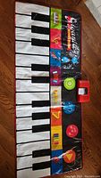 Large kids piano mat laid out on floor showing piano keys and colorful musical instrument icons with an electronic control unit on top right corner.