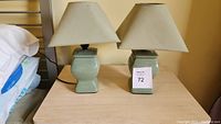 Pair of green ceramic night stand lamps with beige fabric lampshades placed on a light wooden table against a beige wall.