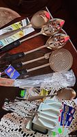 Photo of serving spoons, tongs, ladle, slotted spoons, perforated skimmer, small forks, ceramic condiment spoons, and wrapped plastic utensils on a wooden table with doilies.