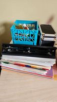 Overview of stationery items including a blue plastic crate full of pens, markers, glue sticks; metal hole punchers; stack of paper and folders.