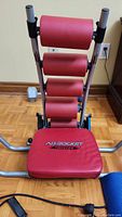 Full front view of Ab Rocket Twister exercise machine showing red padded rollers and seat, gray frame, and part of the yoga mat visible on the floor.