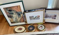 Six framed art pieces arranged on floor: three watercolor cityscape drawings and three small round floral needlepoint pieces framed in gold-tone frames.