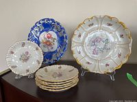 Full set of Dresden china including large white platter, six white side plates with gold trim and floral designs, and a blue decorative platter