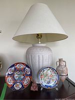 Front view of ceramic lamp with fabric shade, two Asian style decorative plates, decorative urn on stand, and small Buddha figure arranged on surface.