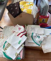 Various linens including white tablecloths, embroidered napkins, crocheted doilies, and colorful decorative mats.