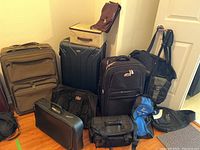 Photo showing multiple luggage pieces including three rolling suitcases and various duffle and handbag style bags arranged on the floor near a wall.